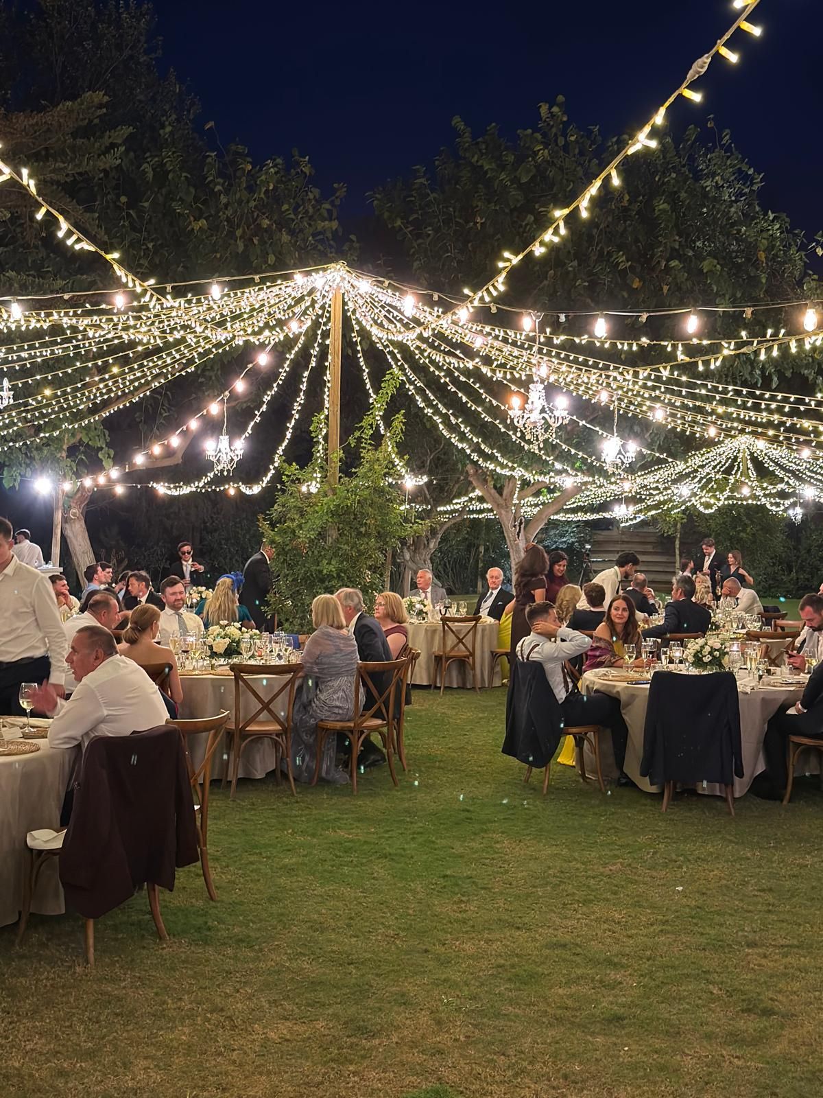 Wedding guests dining outdoors at night under a canopy of warm fairy lights